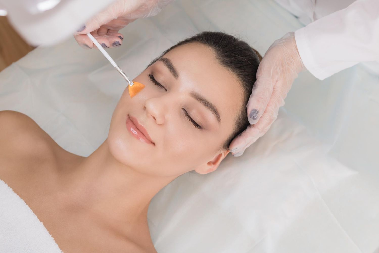 Woman receiving facial treatment, with gloved hands applying product to her cheek.