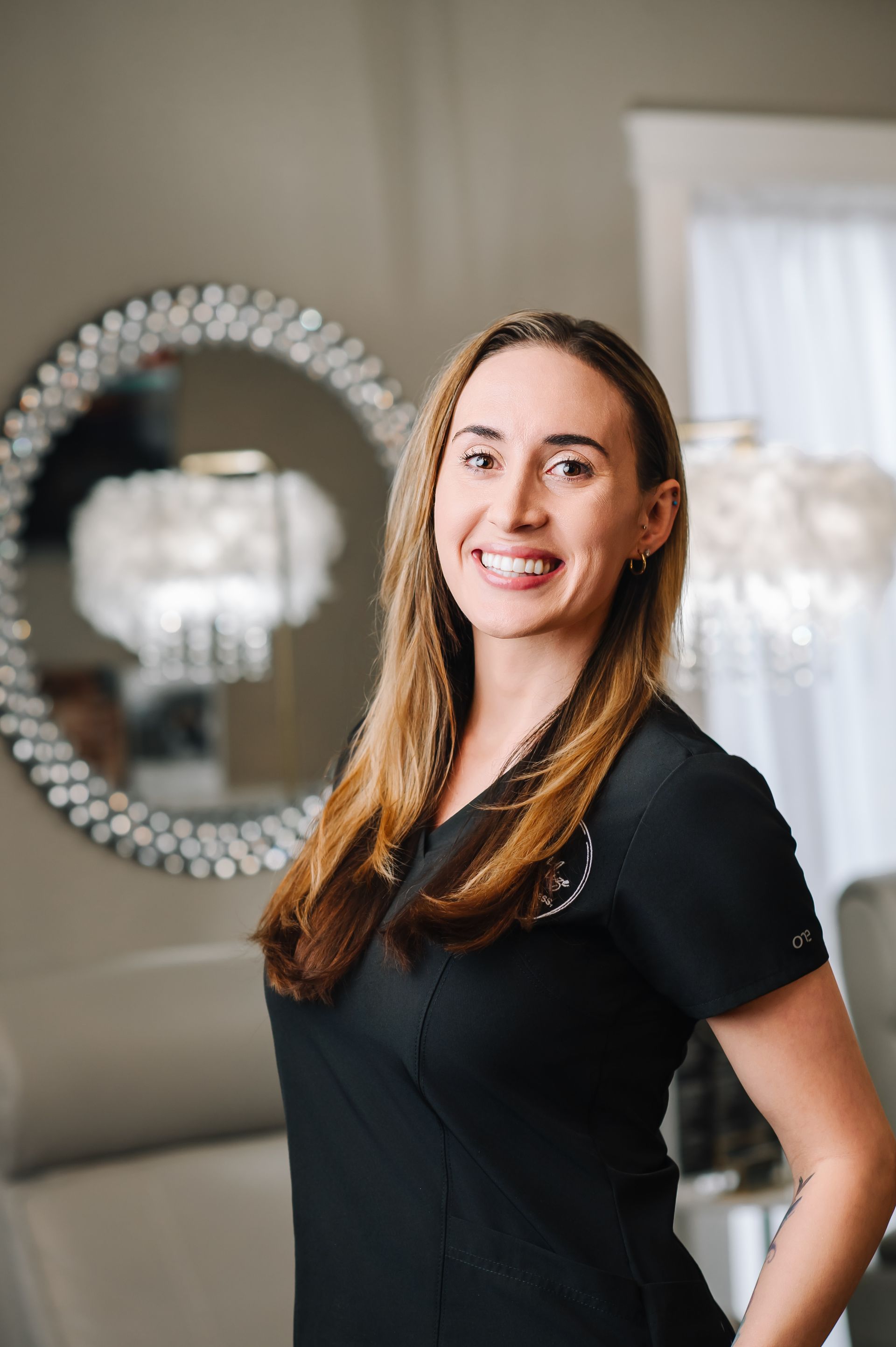 A woman in a black scrub top is smiling and standing in front of a window.