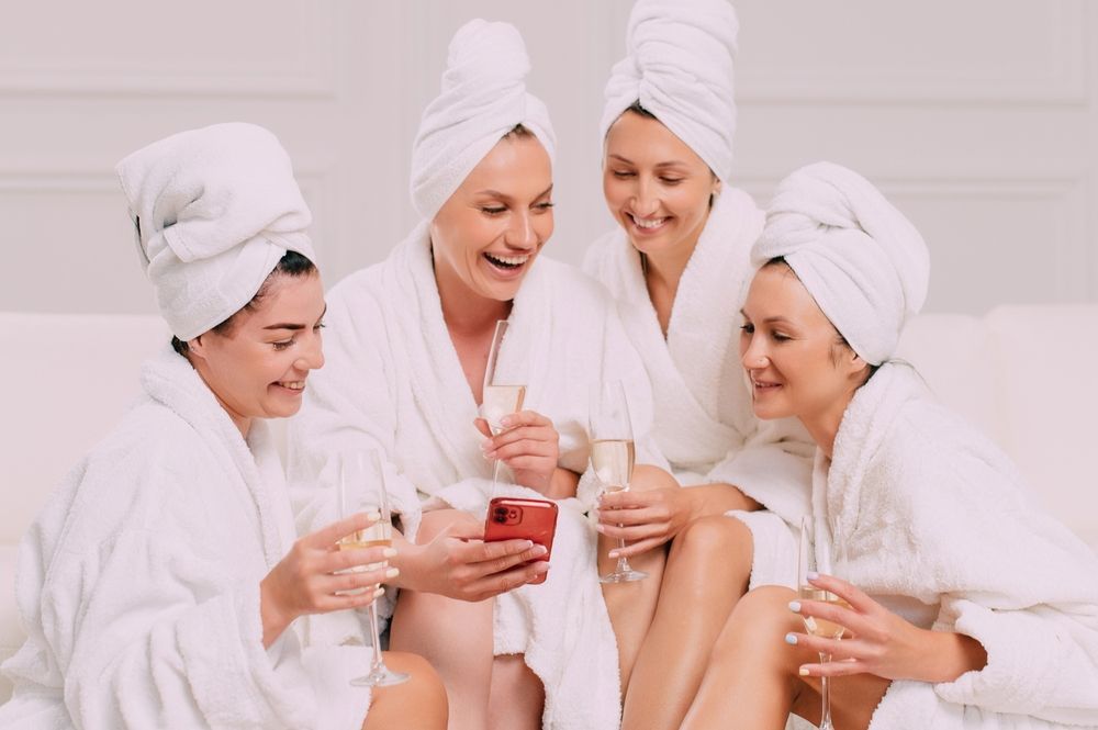 Four women in robes and towels, smiling and looking at a phone together, holding champagne flutes.