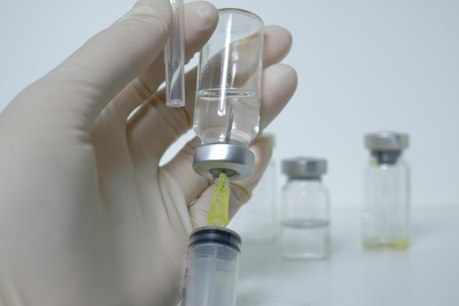 Gloved hand filling a syringe from a vial; other vials are in the background, all on a white surface.