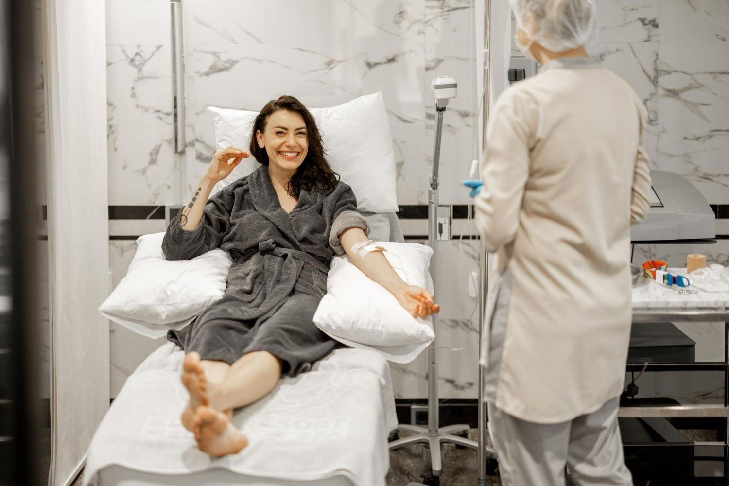 Woman receiving IV drip in a spa-like setting, smiling at camera, medical professional attending.