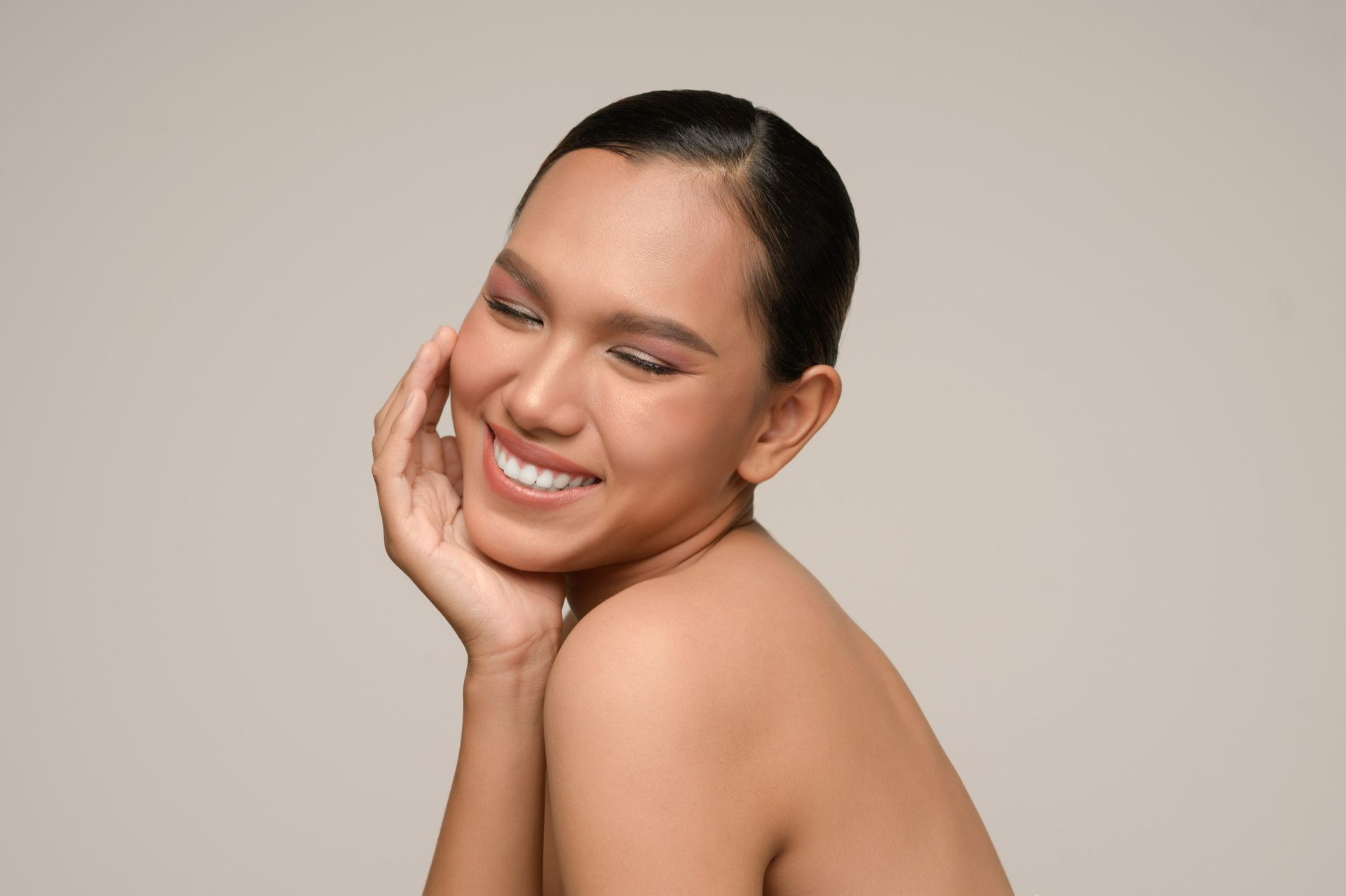 Woman with dark hair and skin smiles, touching her cheek with a hand. Beige background.