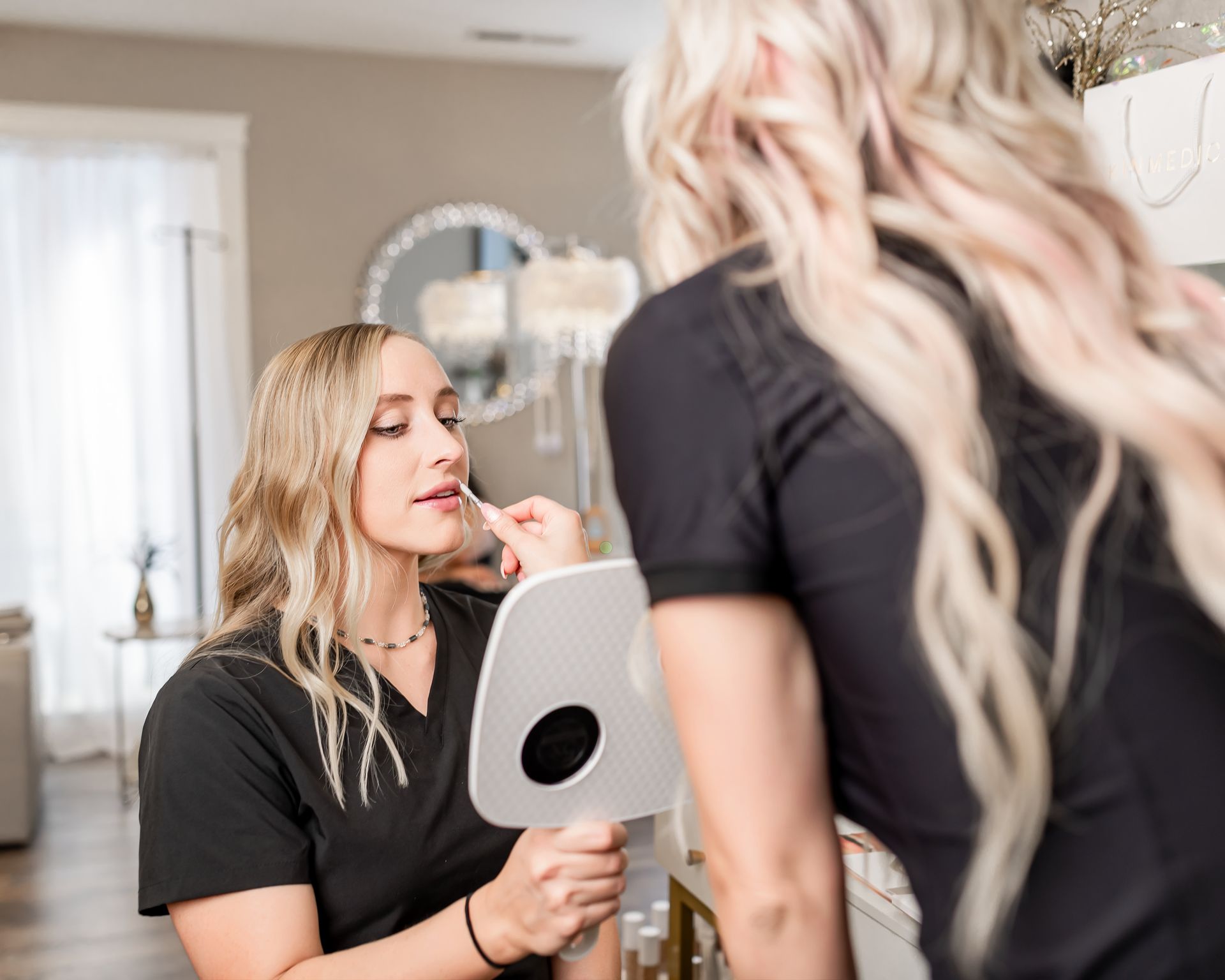 A woman is applying makeup to another woman in front of a mirror.