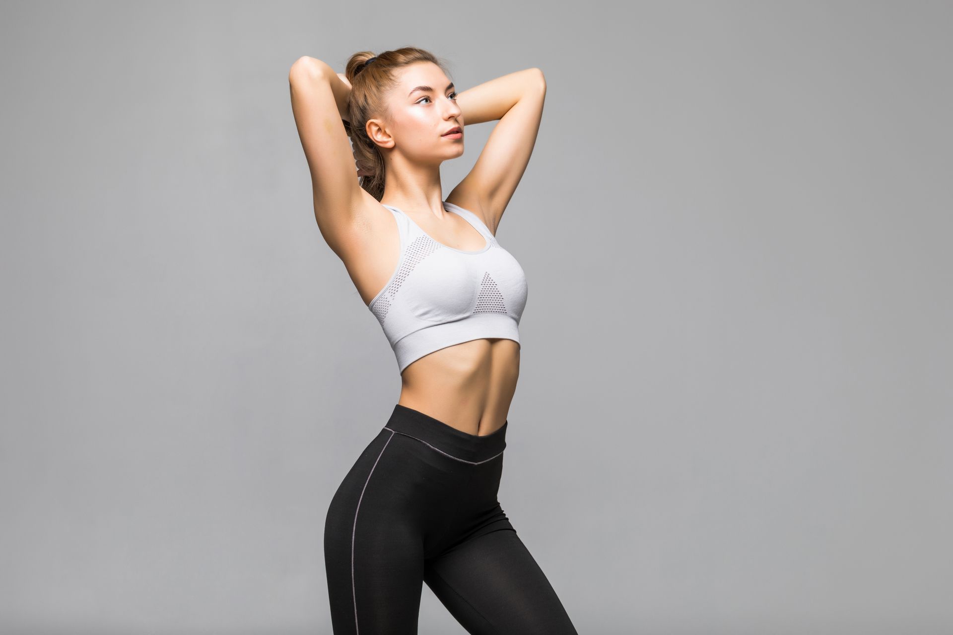 A woman in a white sports bra and black leggings is standing with her hands behind her head.