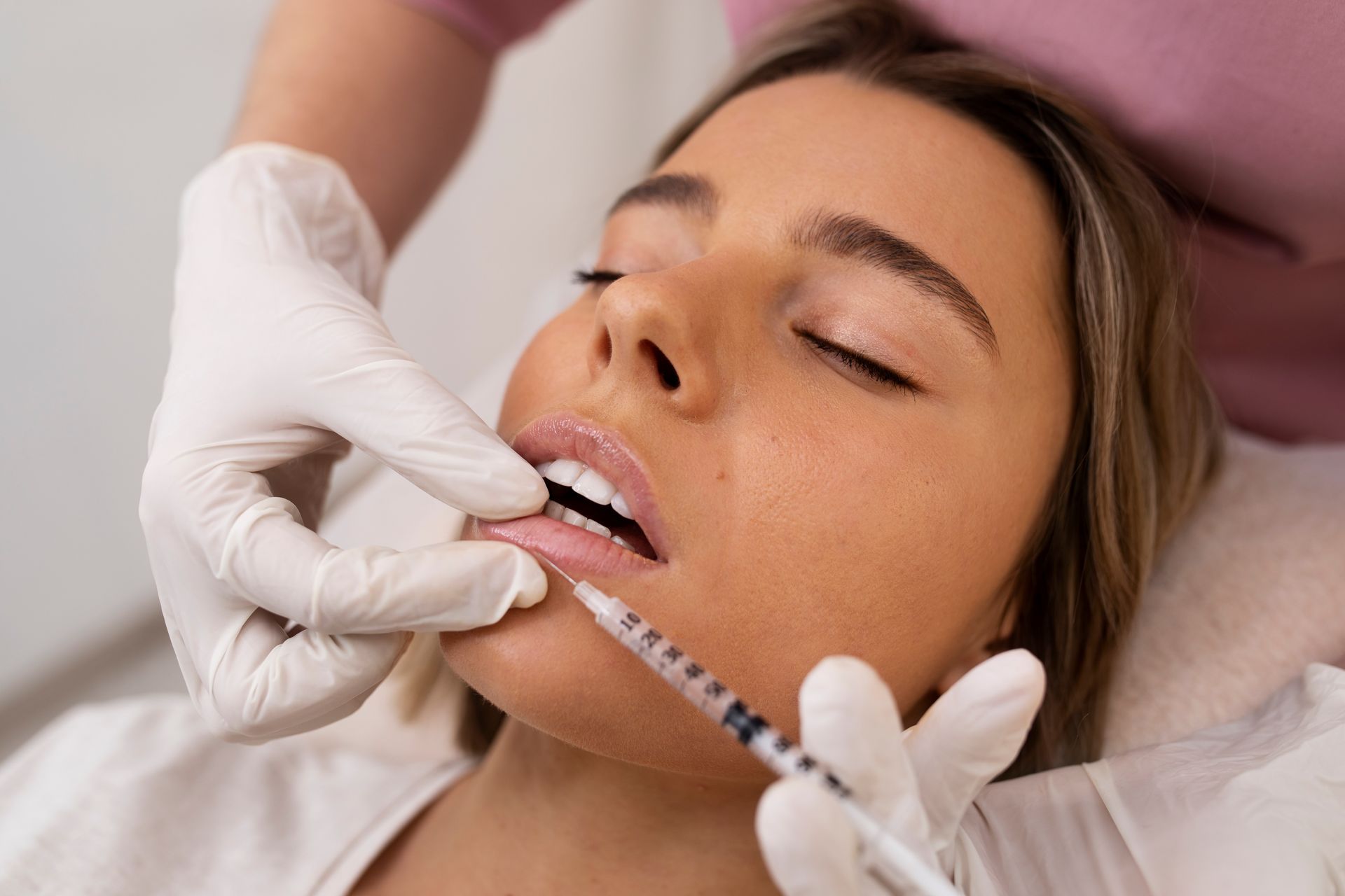 A woman receiving lip filler injections from a gloved hand, eyes closed.