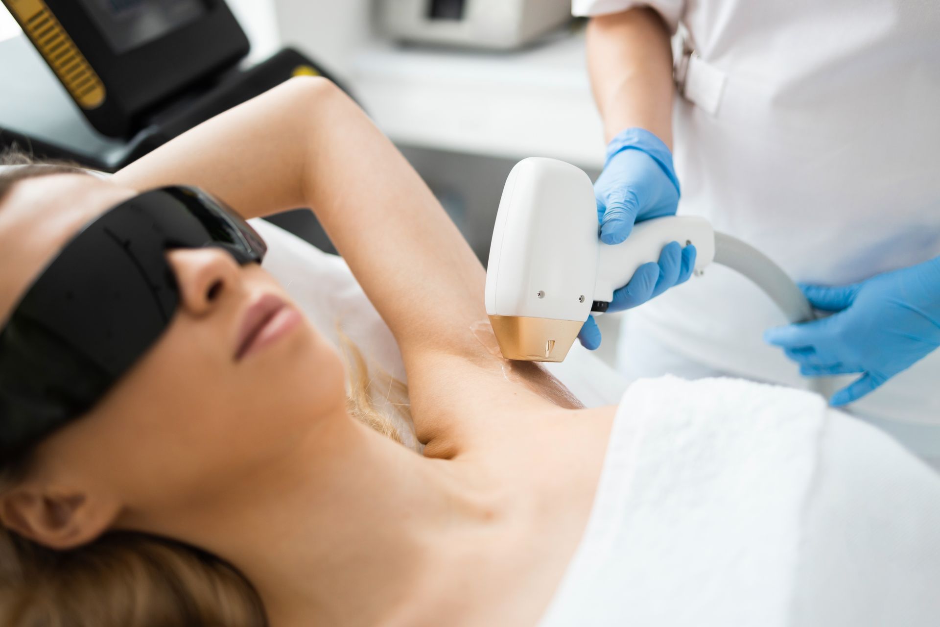 Woman undergoing laser hair removal on her underarm at a clinic; a technician uses a handheld device while the patient wears protective eyewear.