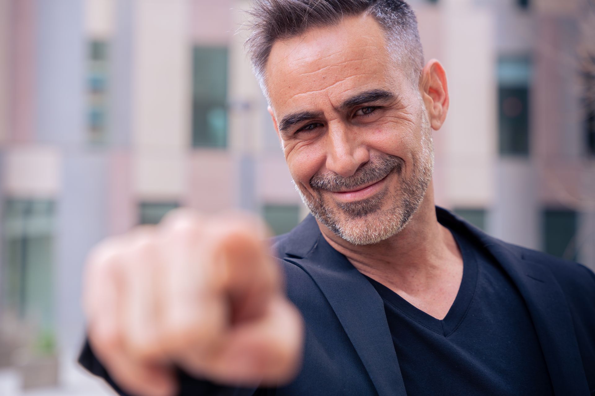 A man in a suit and black shirt is pointing at the camera.