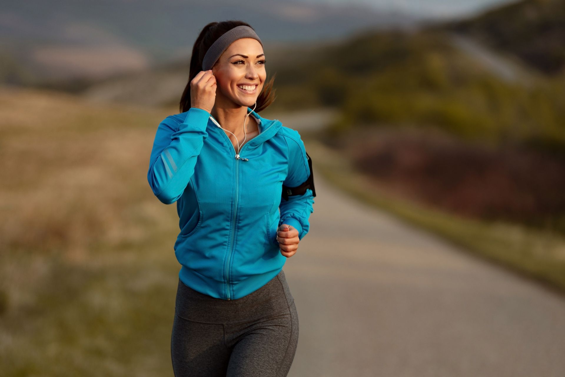 Woman jogging on a paved road in nature, wearing a blue jacket, gray leggings, and a headband; smiling and looking to the side.