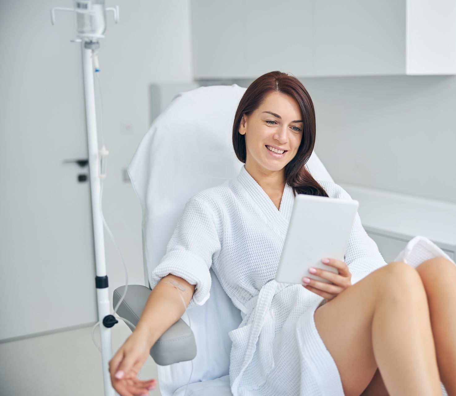 A woman in a bathrobe is sitting in a hospital chair holding a tablet.