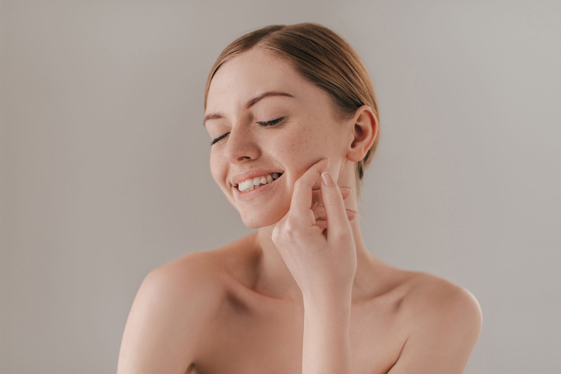 A fair-skinned woman smiles with closed eyes, touching her cheek. She has freckles, and a neutral background.