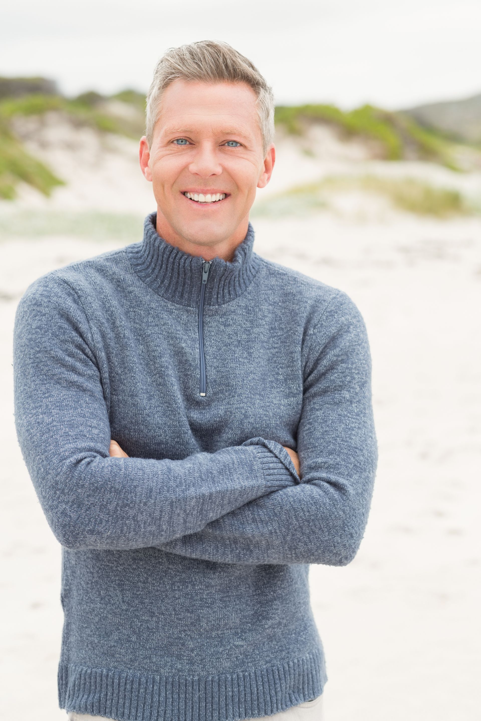 A man in a blue sweater is standing on the beach with his arms crossed and smiling.