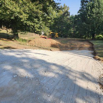 A gravel driveway leading to a construction site with trees and a yellow excavator in the background.