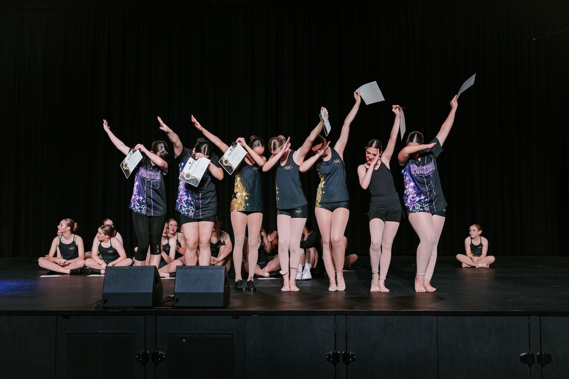 Dancers on stage with arms raised. Some wear t-shirts and shorts. Dark lighting.