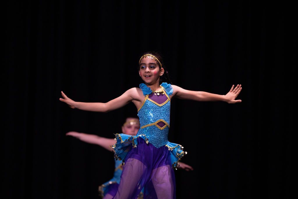 Dancers on stage, arms outstretched. The lead dancer in blue and purple costume smiles with a jeweled headband. Dark background.