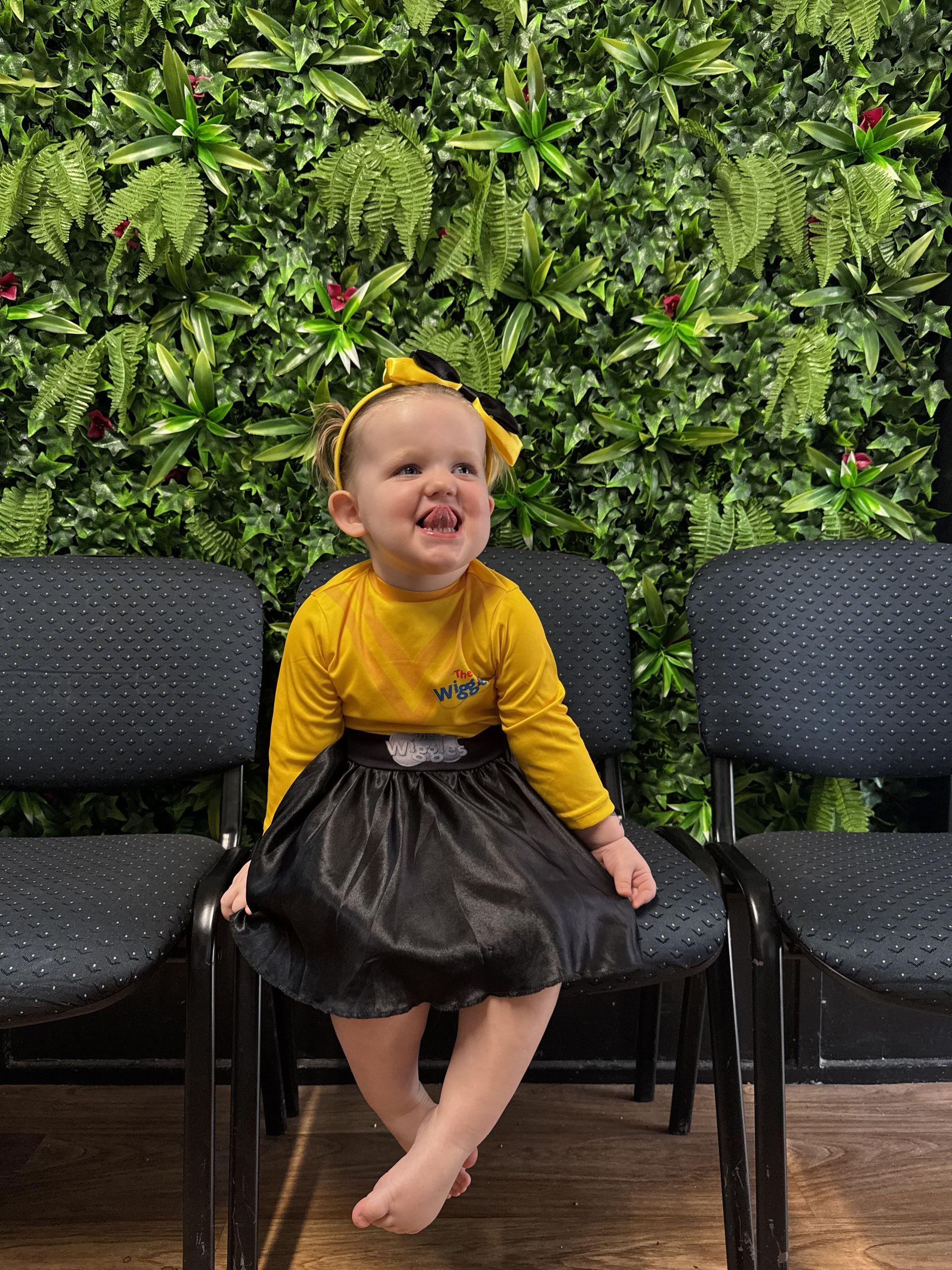 Smiling child with yellow top and black skirt sits on a chair in front of a green wall.