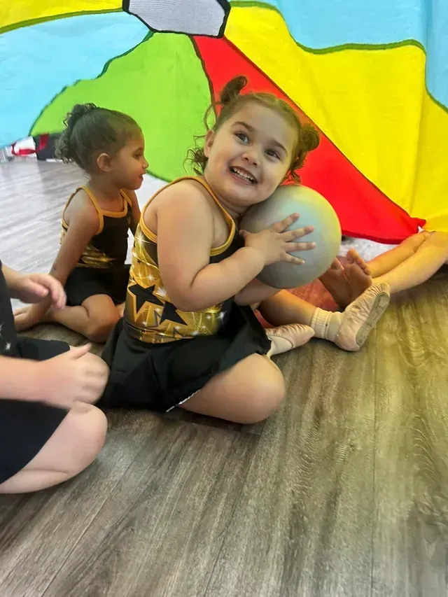 Young girl in colorful tutu and bow, posing on stage with hands under her chin.