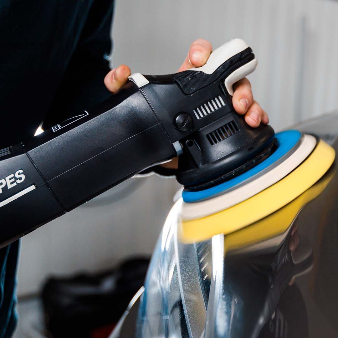 A person polishing a car with a black and white polisher. The polisher has a yellow, blue, and white pad.