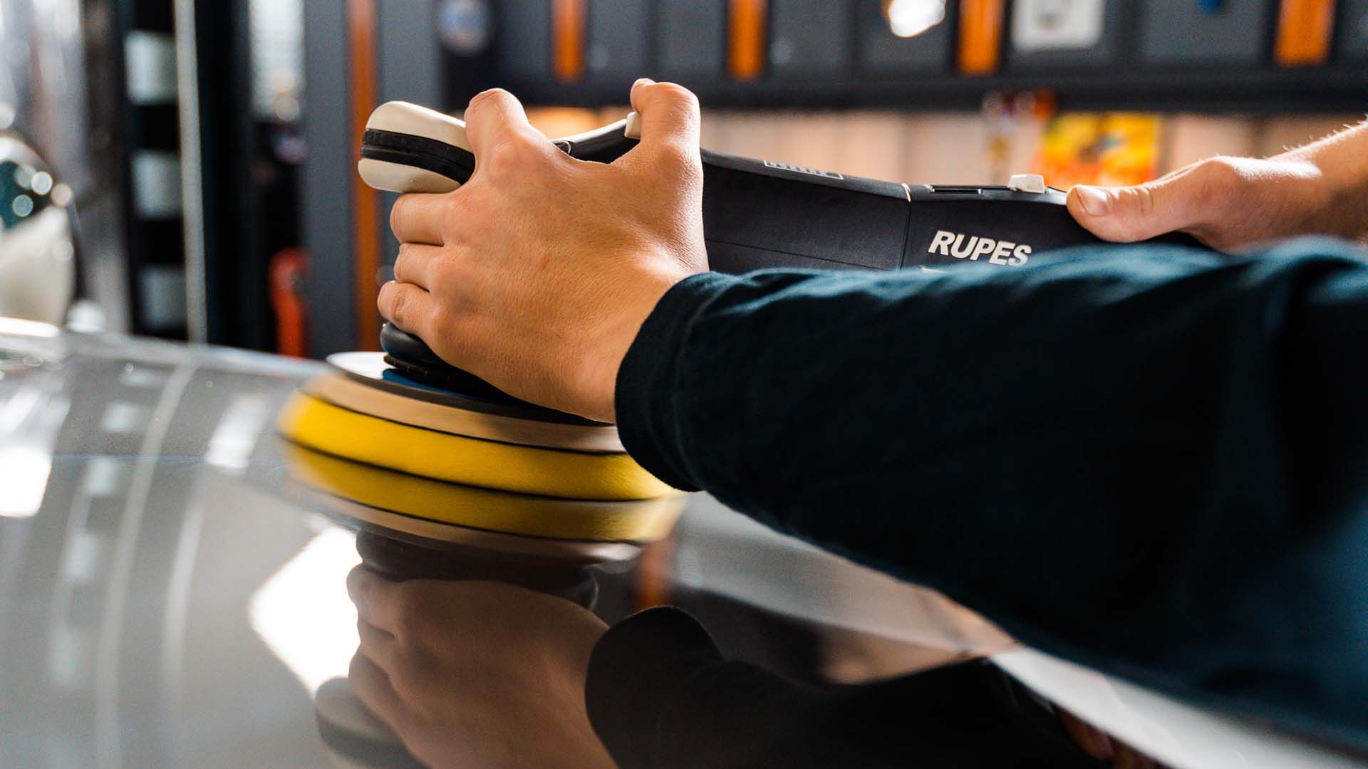 Hands using a Rupes polisher on a car's surface, likely buffing or detailing in a garage.