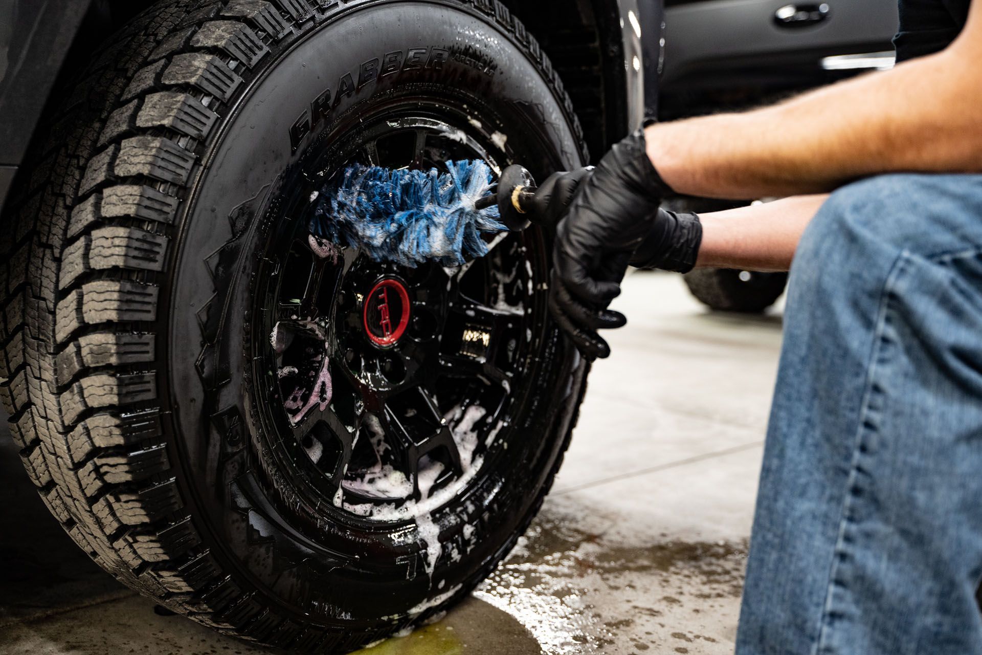 Person washing a black car wheel with a soapy rag, wearing black gloves and blue jeans, in a garage.