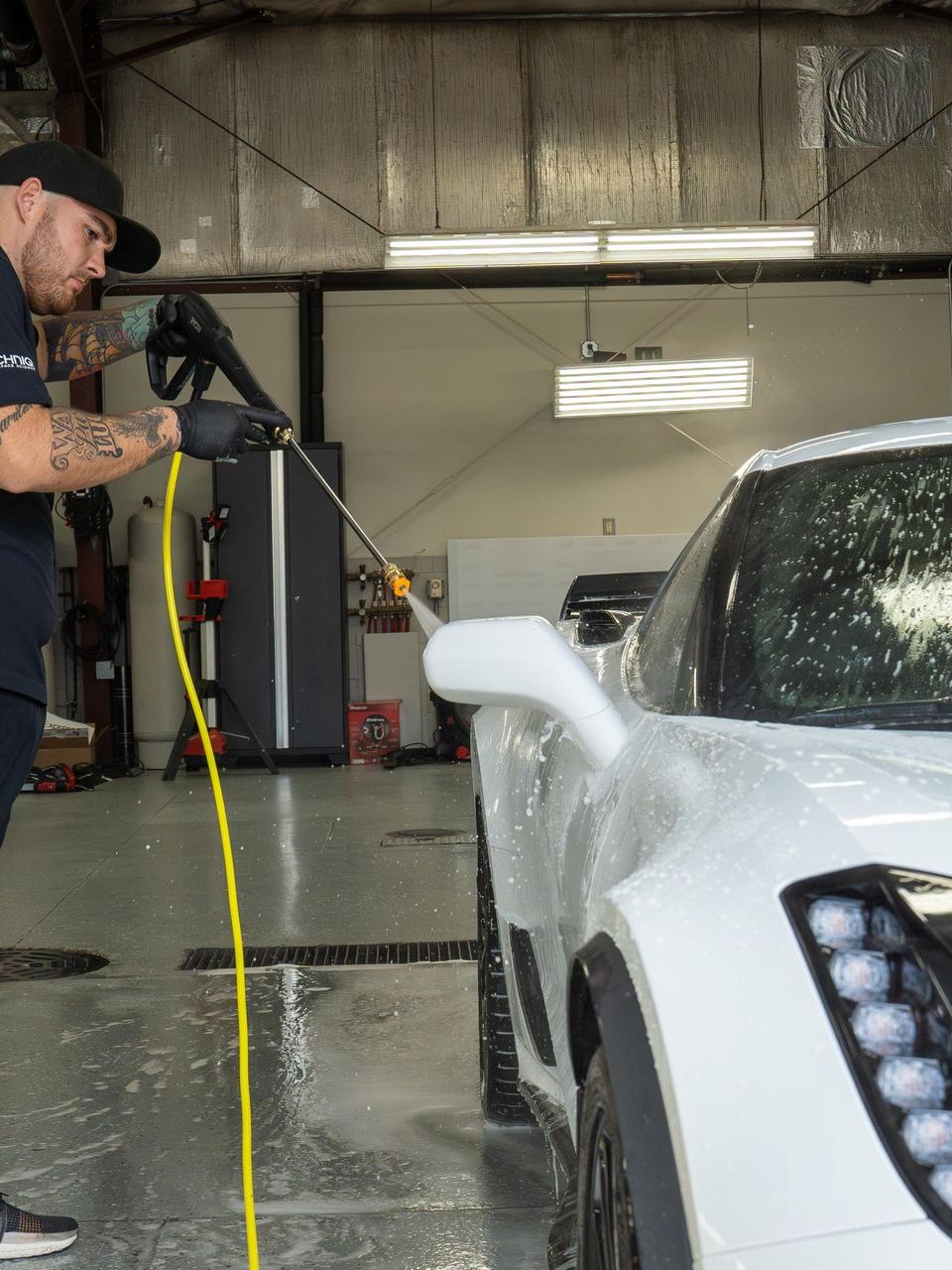 Man washing a white sports car with a pressure washer inside a garage.