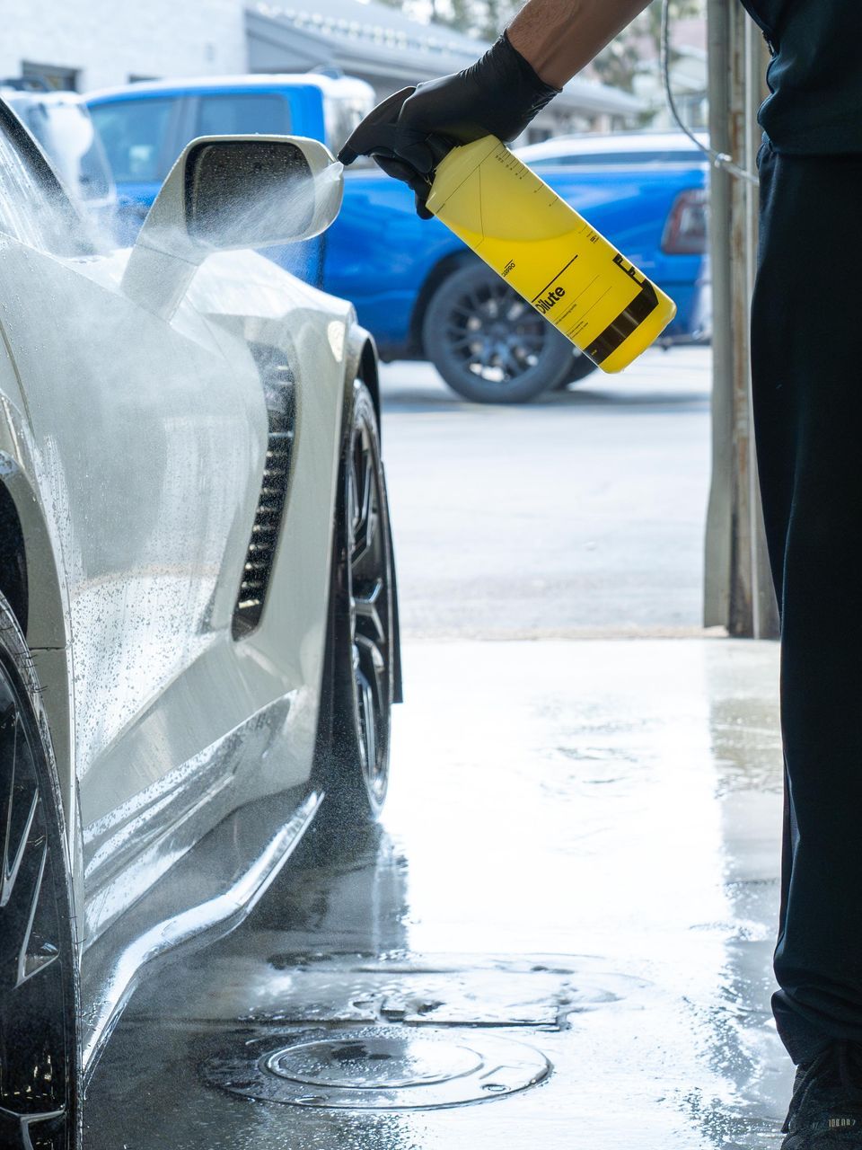 Person washing a white car with soap using a yellow spray bottle at a car wash. The car is parked with water and soap suds all over it.