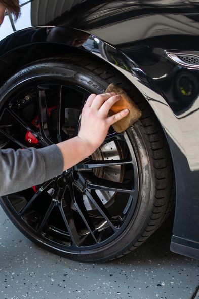 Person washing a black car tire with a sponge, with the vehicle's shiny black body and glossy black wheel visible.