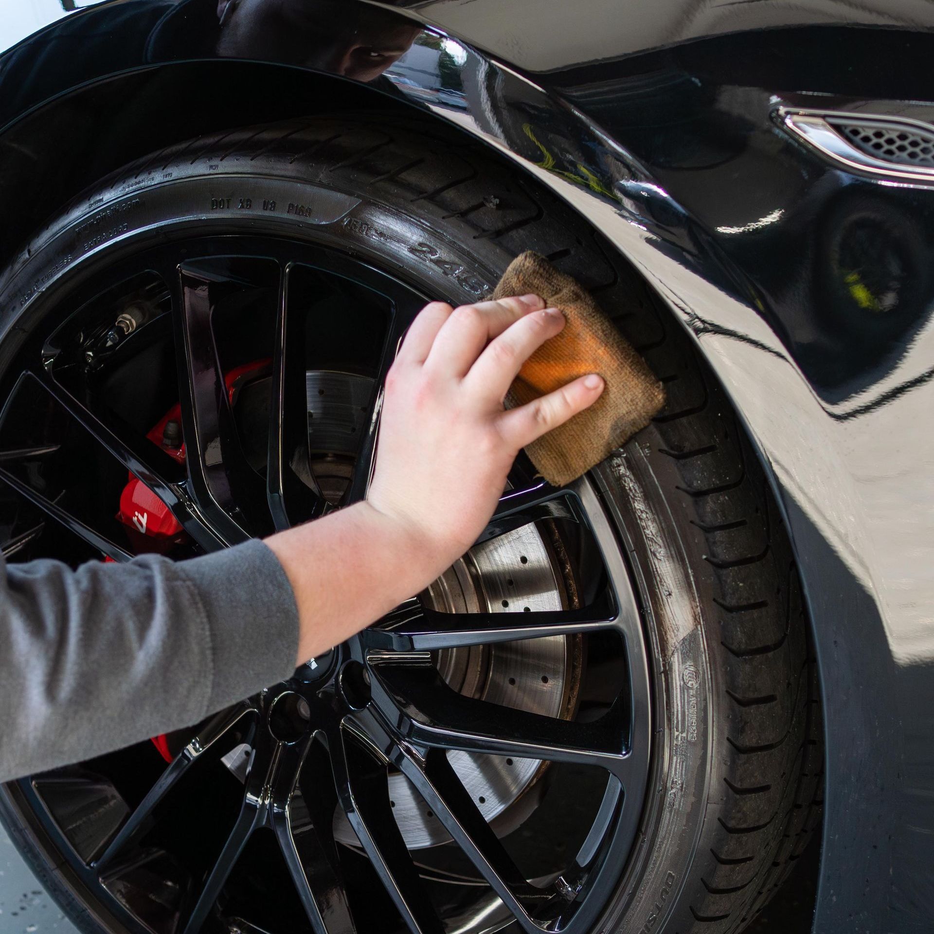 Person's hand with a sponge cleaning a black car tire. The car has a shiny black finish and red brake calipers.
