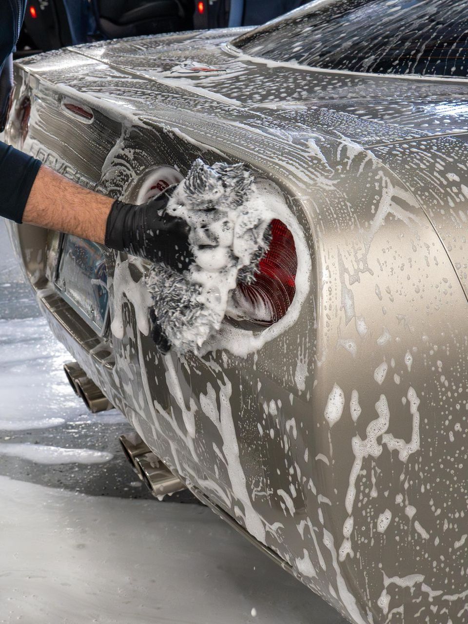 Person washing a silver car's rear with a soapy cloth, focusing on a taillight. Black gloves are worn.