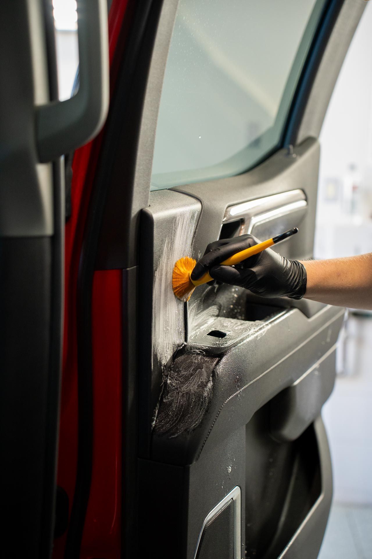 A person wearing a black glove cleans a car door panel with a brush. Red car door is open in a garage.