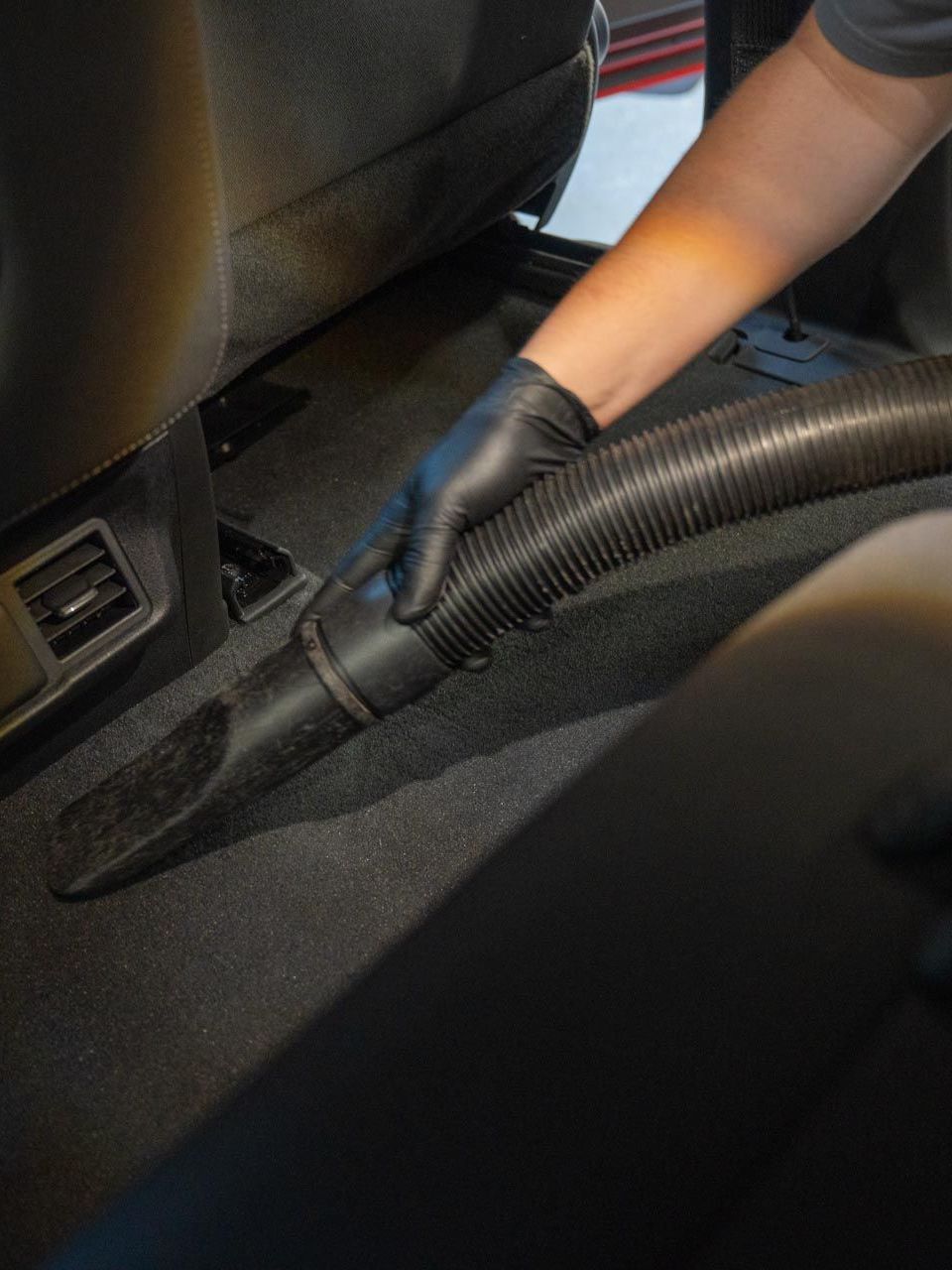 A person wearing black gloves vacuums a car interior with a hose, focusing on the carpet.