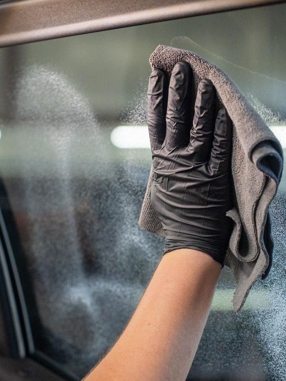A person wearing a black glove wipes a car window with a gray microfiber cloth, removing spray.