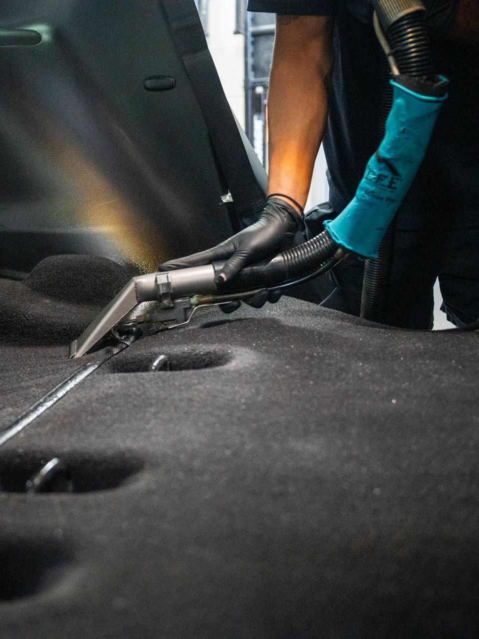 A person in black gloves and a blue sleeve vacuums a car's black carpet.