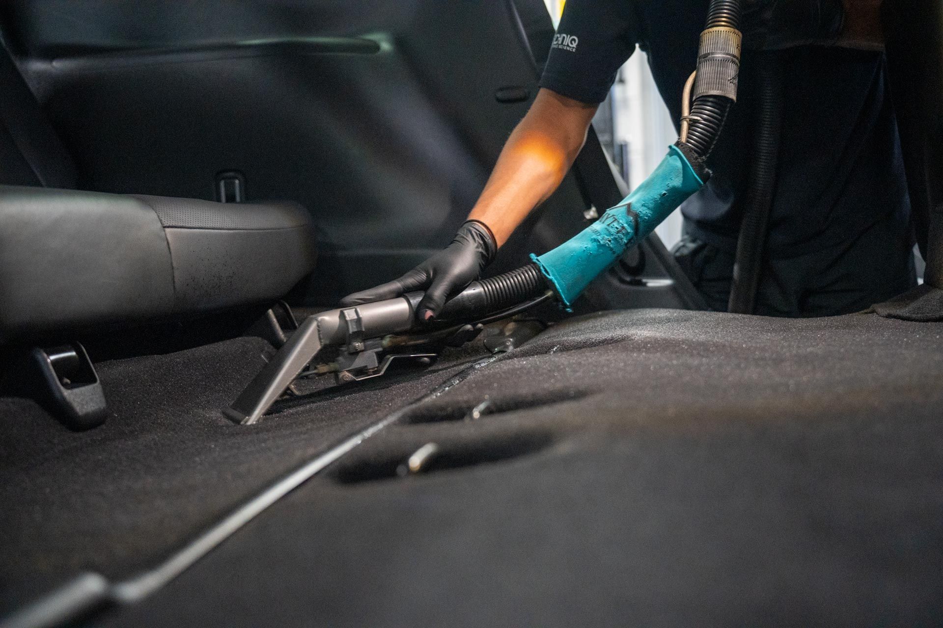 Person wearing gloves vacuums a car's carpet with a handheld machine. The setting is the inside of a car.