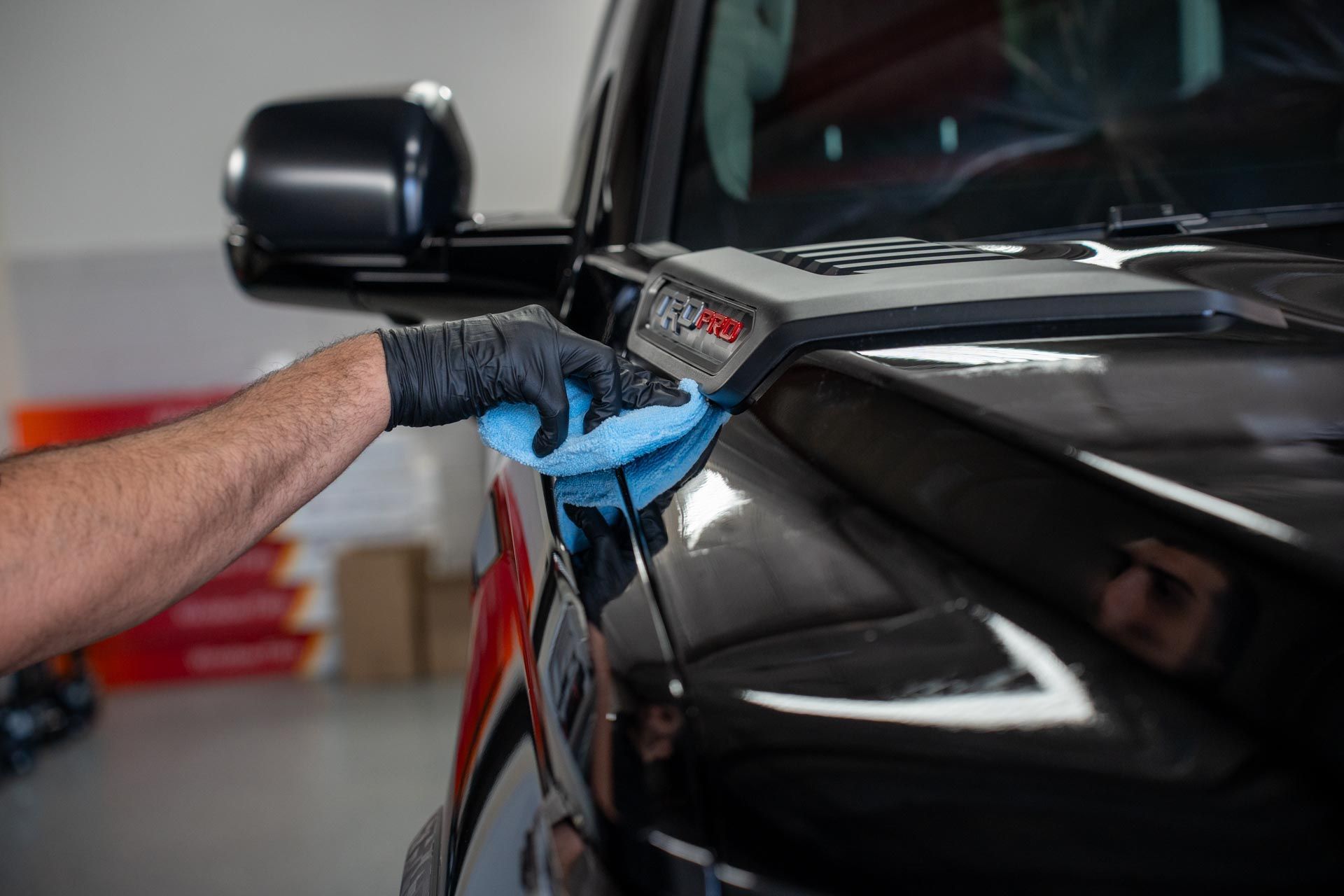 A person wearing a black glove wipes a shiny black car hood with a blue cloth. Inside a garage.
