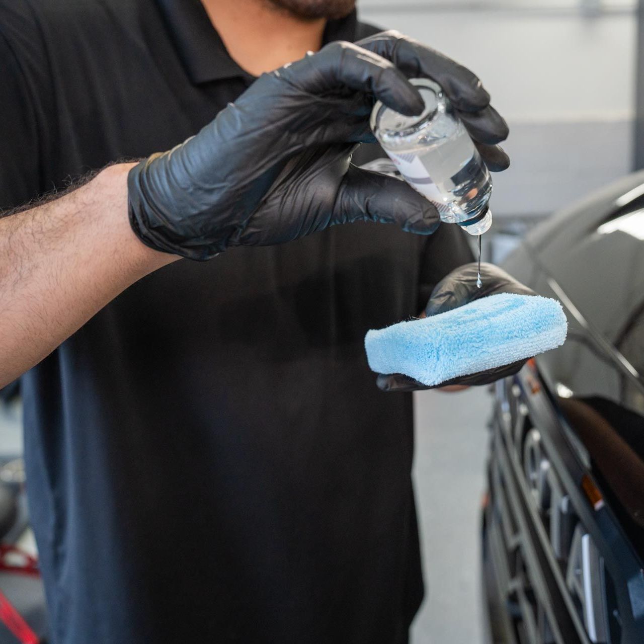 A person wearing black gloves applies a liquid from a bottle onto a blue sponge, working on a black car.