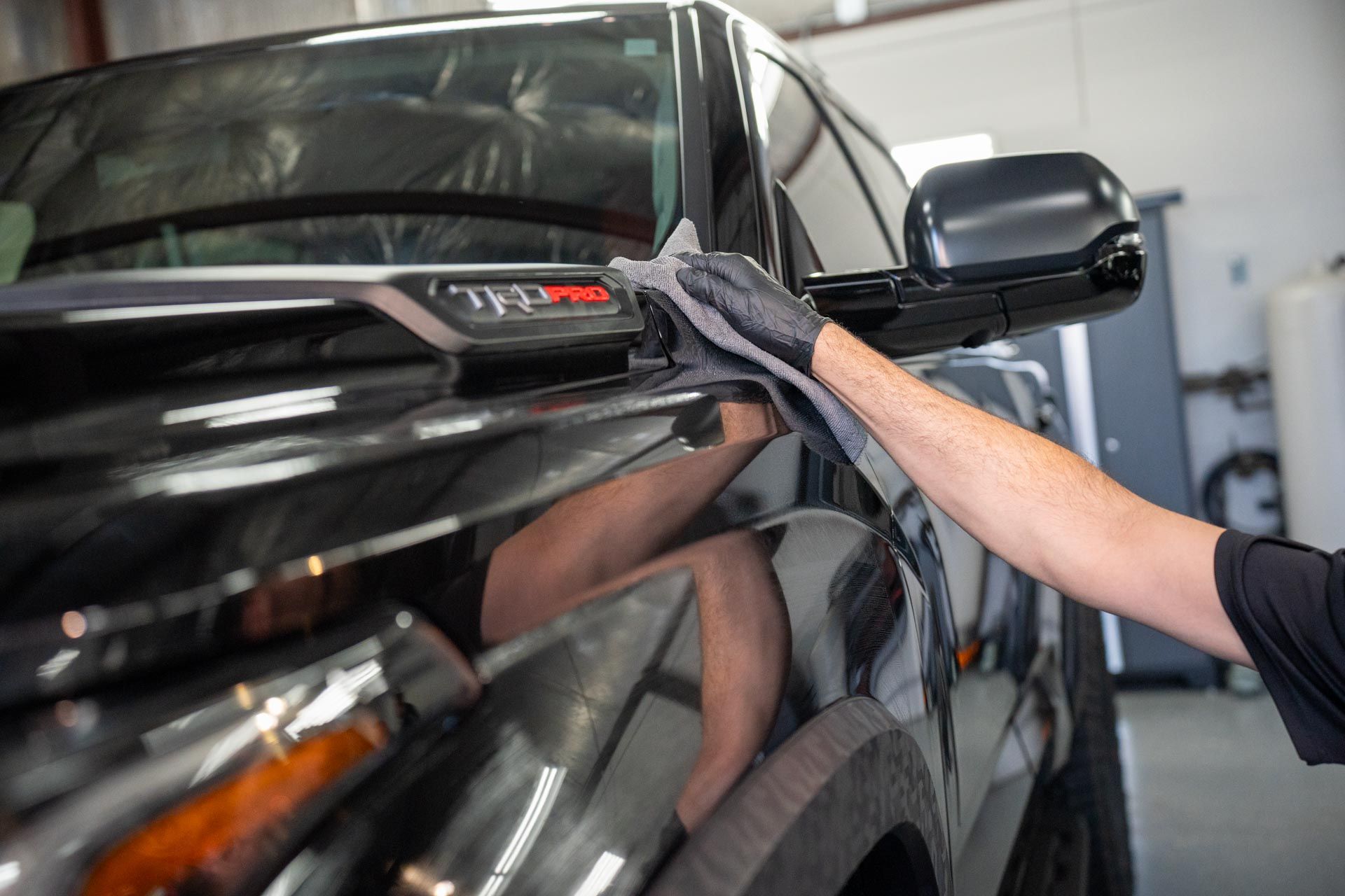 Person wearing black gloves wiping a black truck with a gray cloth; detail shot in a garage.