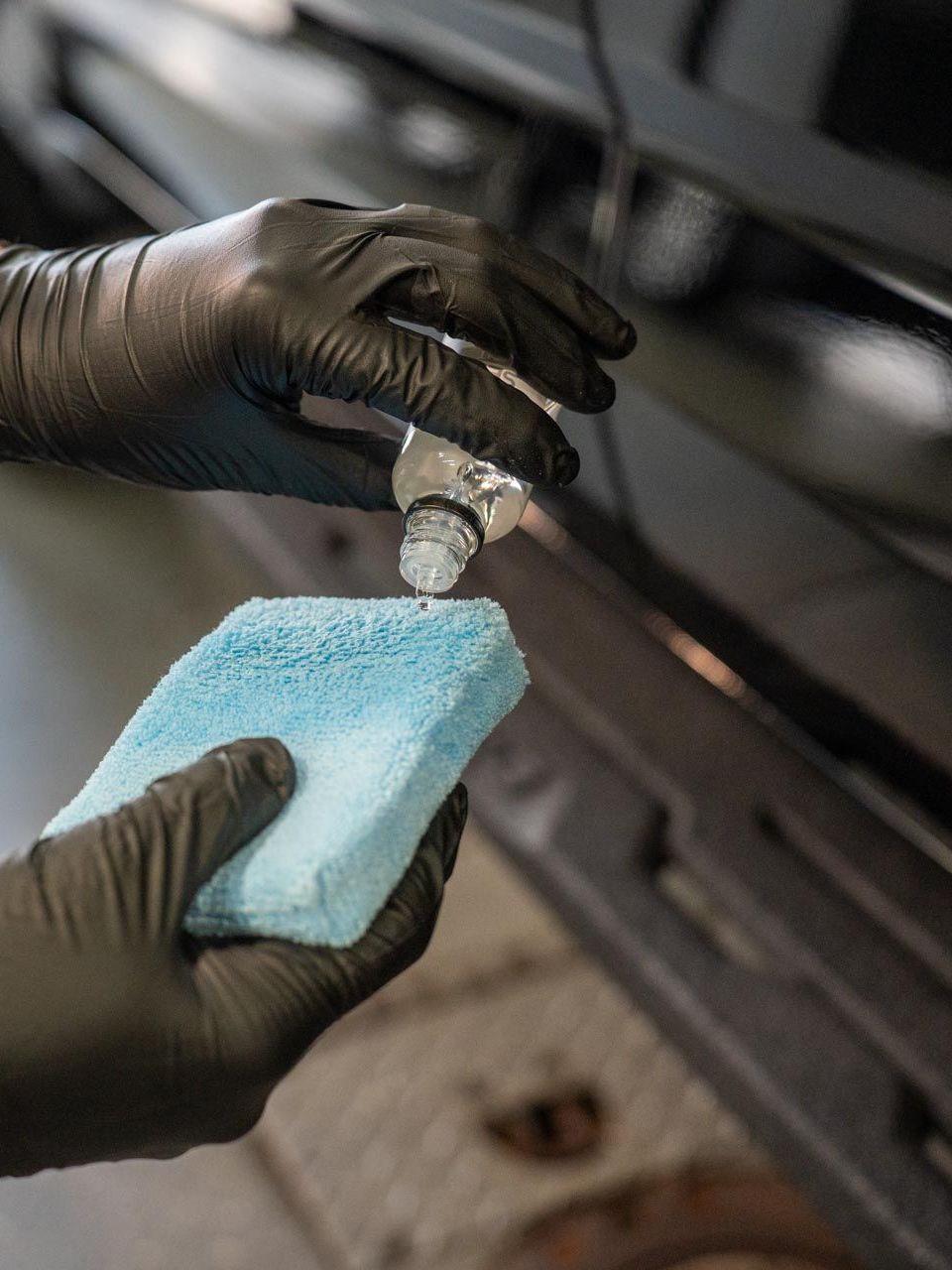 Person wearing black gloves applying liquid from a bottle onto a blue sponge, likely detailing a black car.