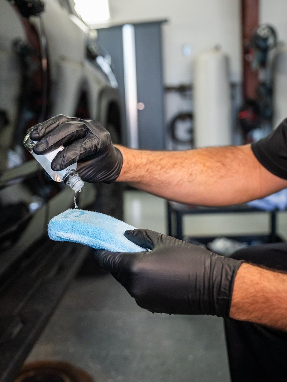 Person in black gloves applying liquid from a bottle onto a blue applicator, next to a black car in a garage.