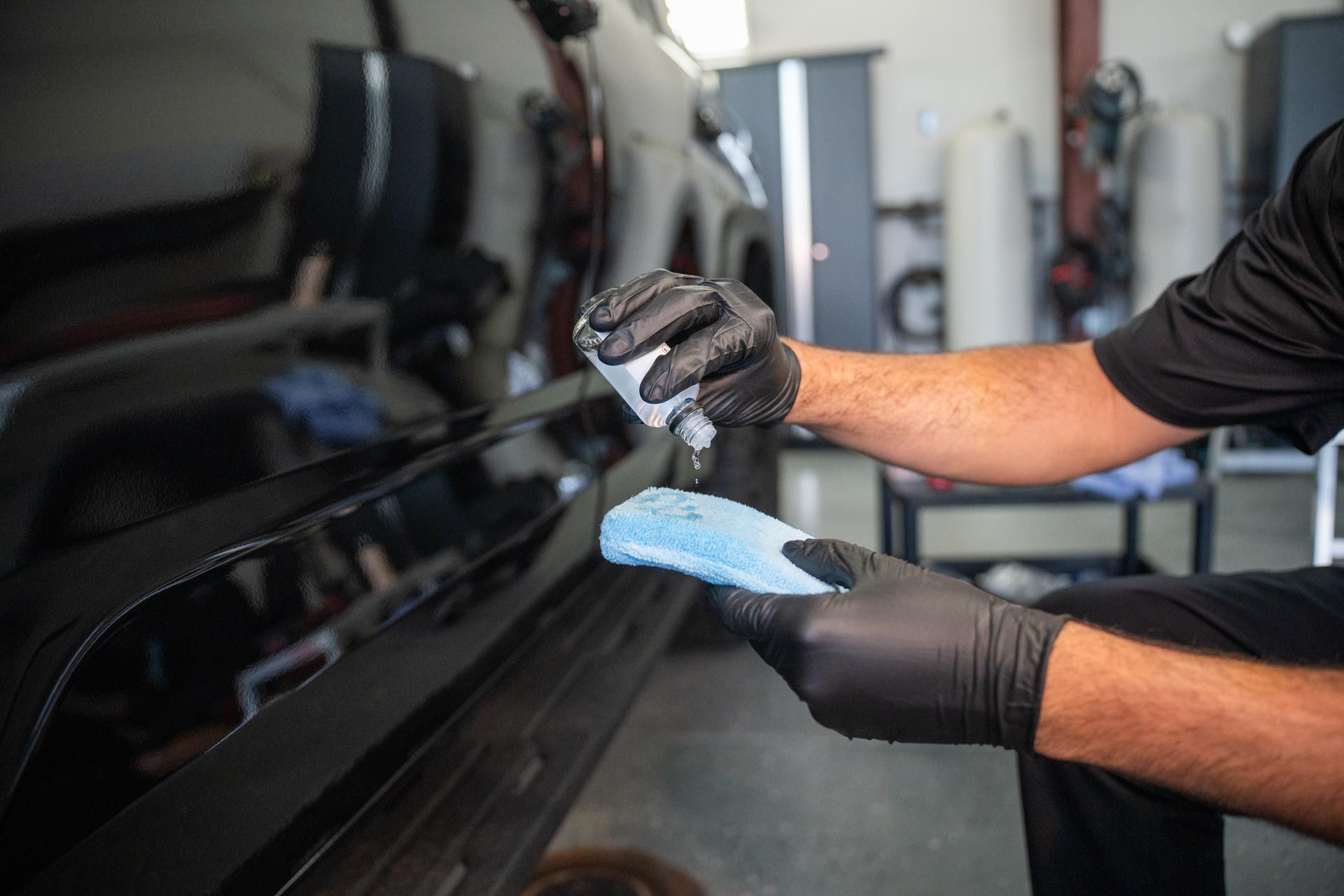 Person applying ceramic coating to a black car with a blue applicator and wearing black gloves. Inside a garage.
