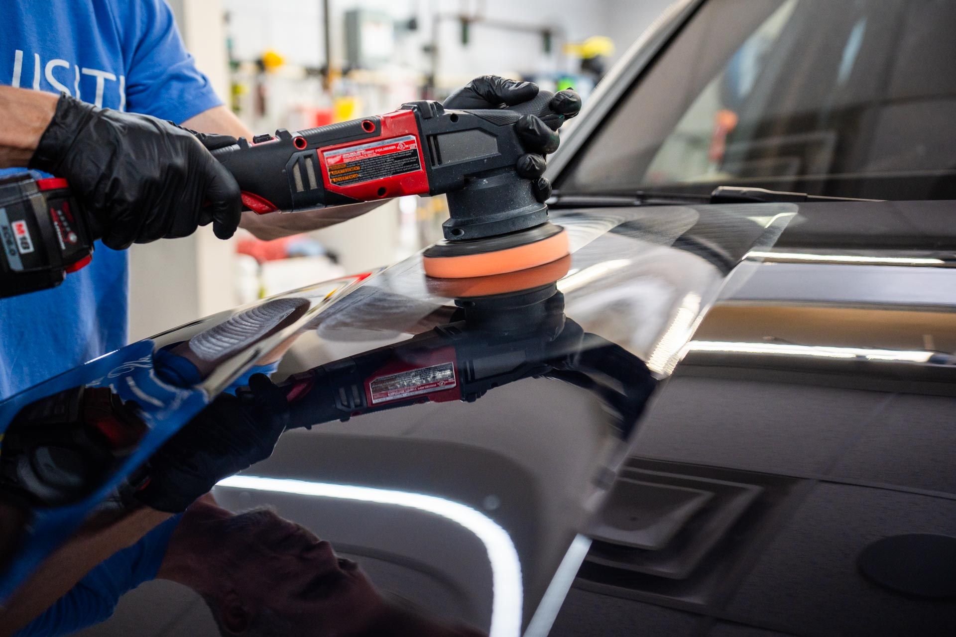 Person in black gloves using a red and black buffer on a black car in a well-lit shop.
