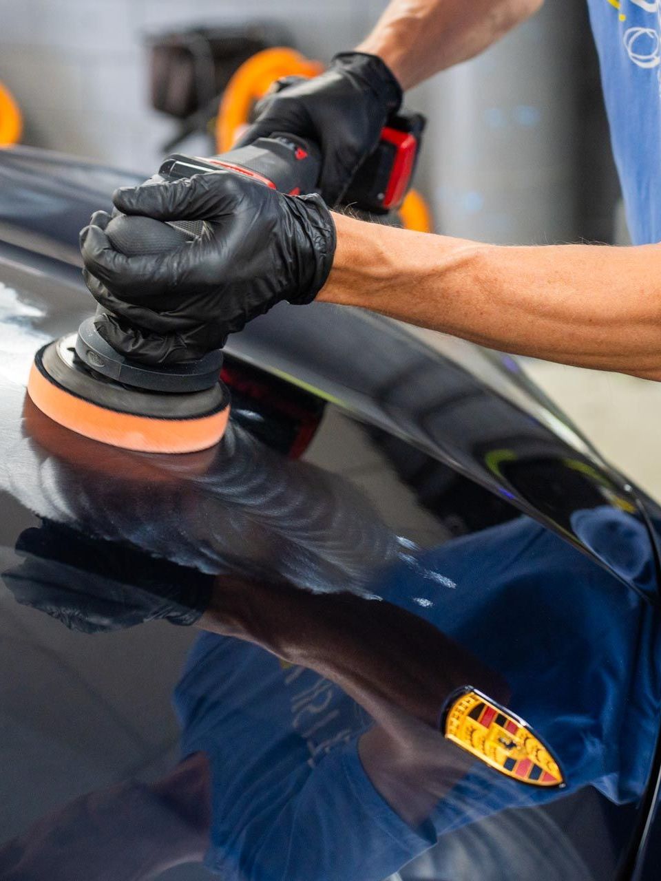 Person in black gloves using a polisher on a black car's hood, next to a Porsche logo.