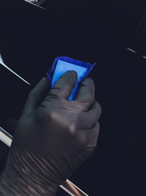 Gloved hand applying a blue detailing clay bar to a black car's surface.