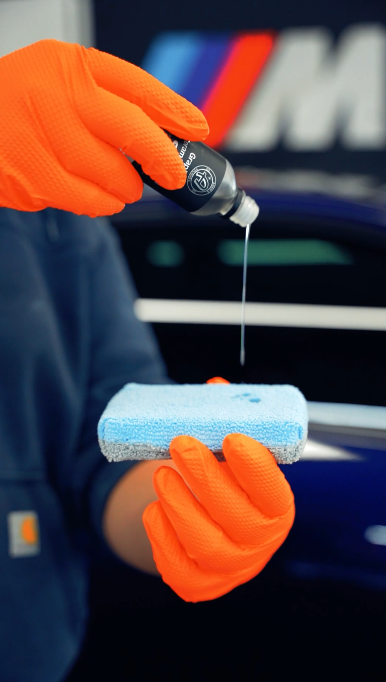 Gloved hands pouring liquid onto a blue foam applicator; a blue car is visible in the background.
