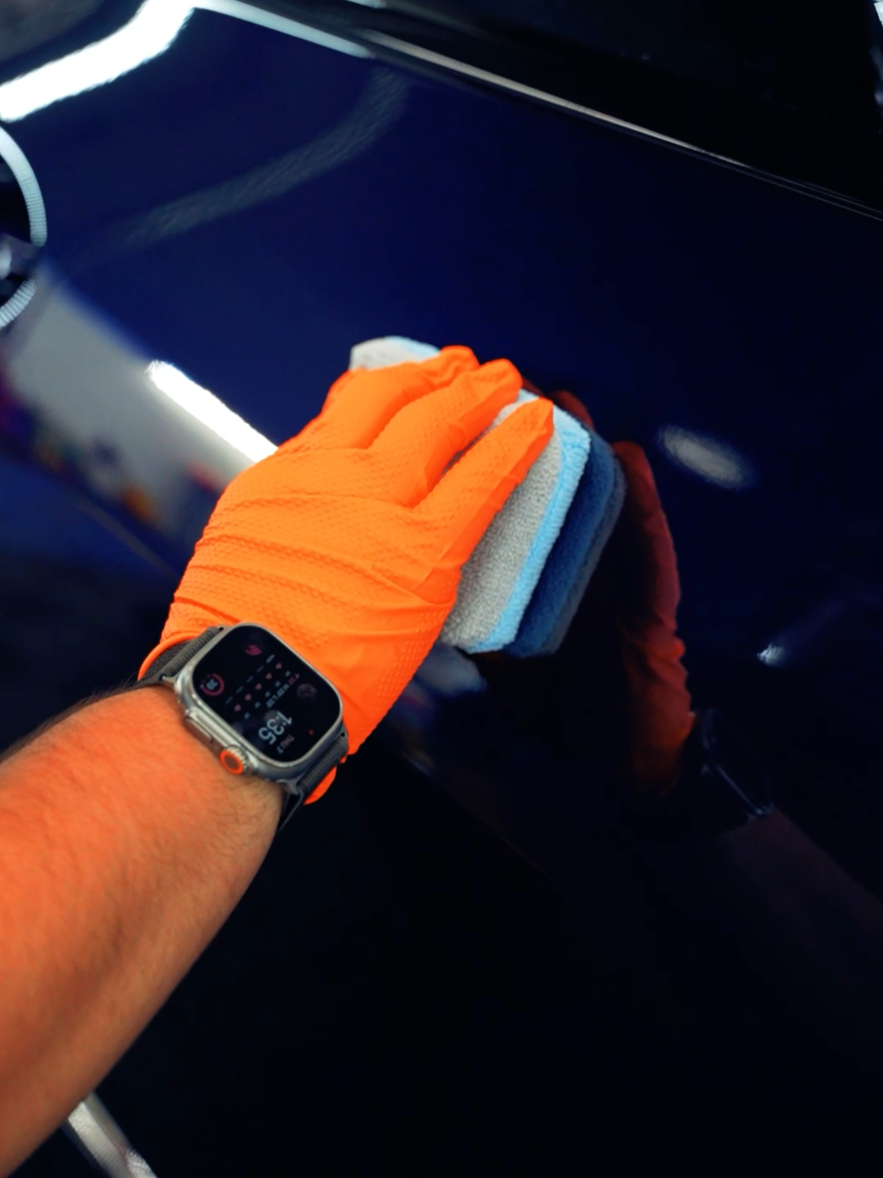 A person wearing orange gloves and a smartwatch applies a blue coating to a dark car surface with an applicator pad.