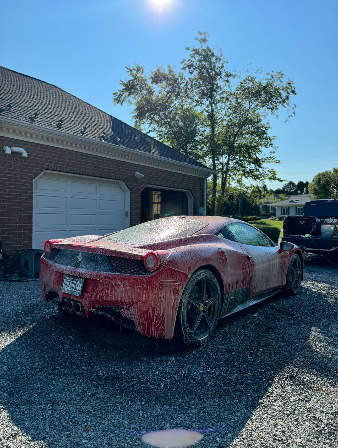Red Ferrari covered in foam, being washed in a driveway with a brick garage on a sunny day.