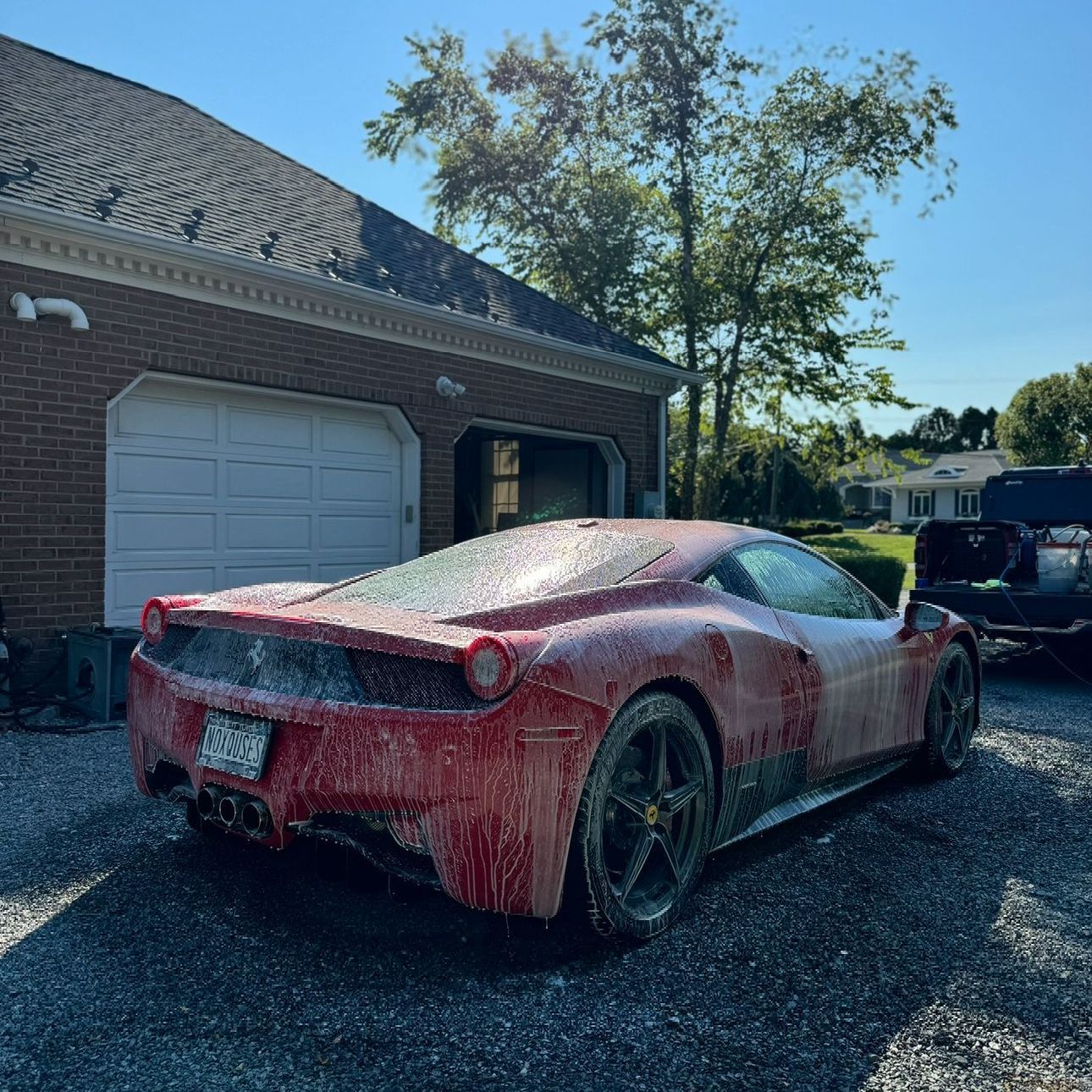 Red Ferrari being washed with soap in a gravel driveway next to a brick garage.
