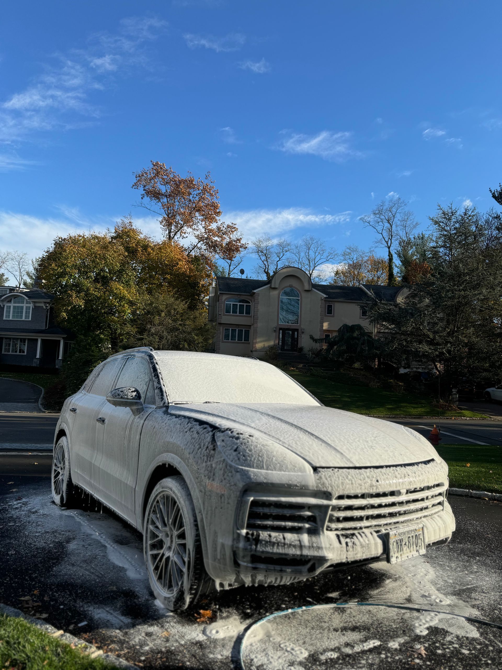 A white SUV covered in foamy car wash soap, parked on a paved surface in front of a house and trees on a sunny day.