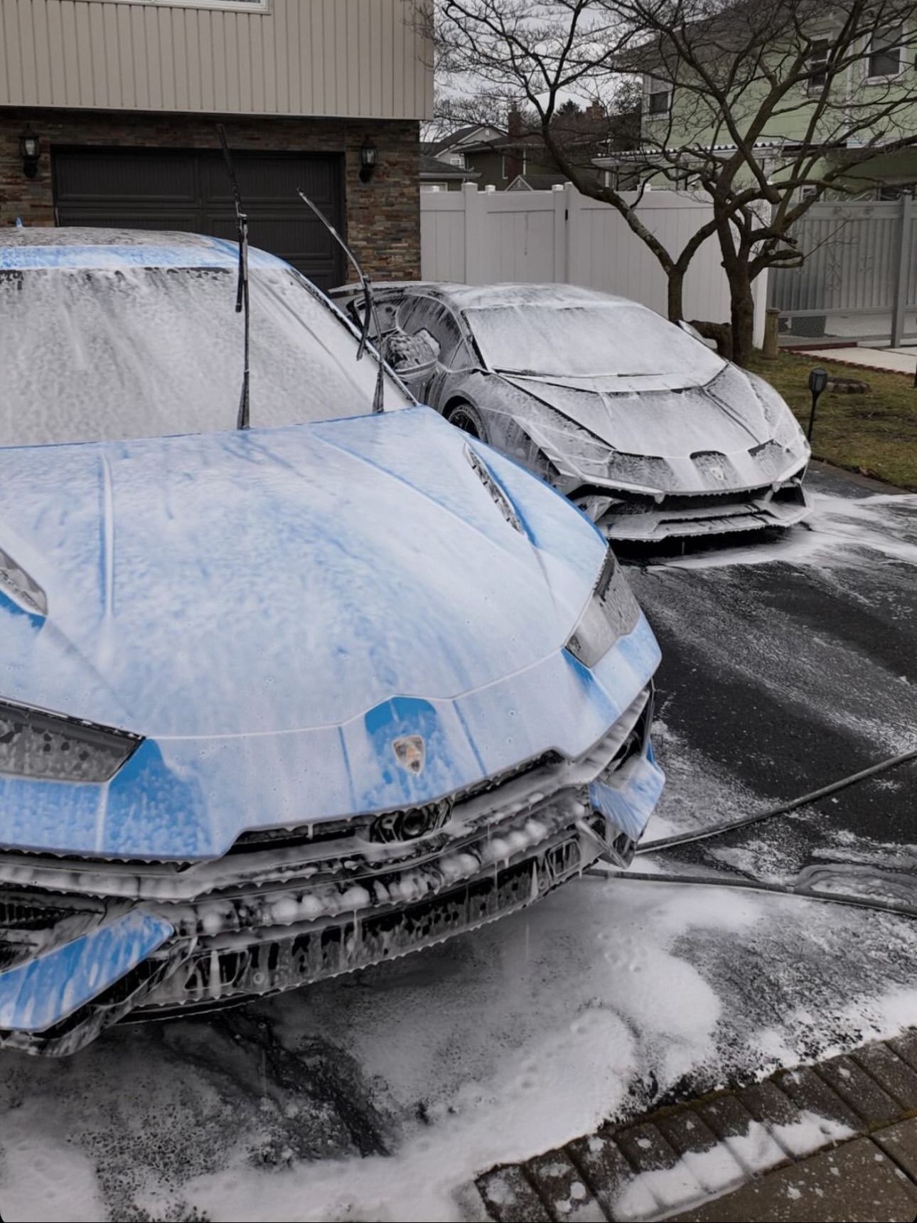 Two blue and grey Lamborghini sports cars covered in foam suds being washed outside a house.