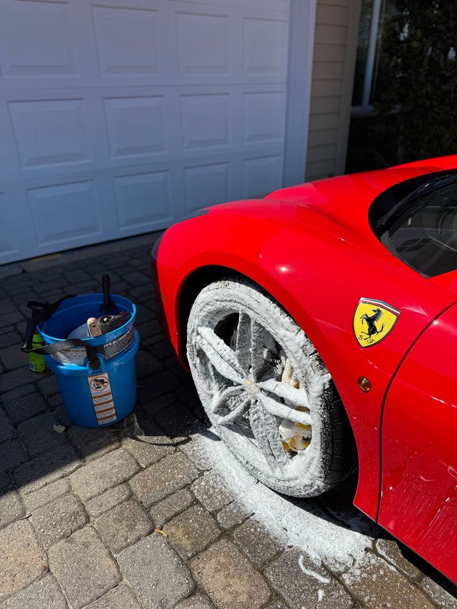 Red Ferrari being washed, with suds on the wheel and a bucket of cleaning supplies nearby on a brick driveway.