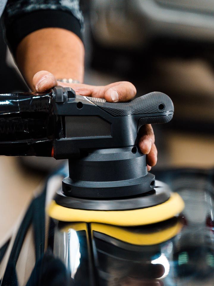 Person using a black car polisher on a vehicle. The polisher has a yellow pad and reflects the car's surface.